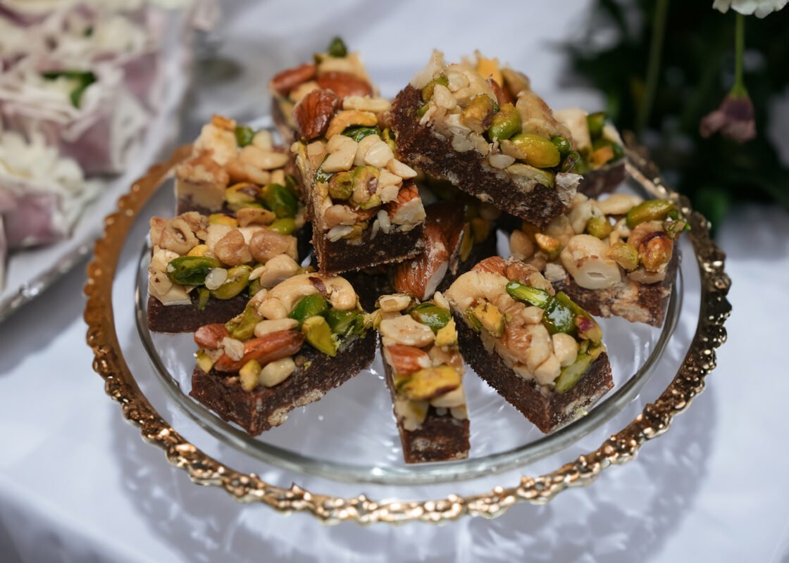 Tamreyeh dessert featuring a soft date dough infused with cardamom, topped with a mix of pistachios, cashews, walnuts, and almonds, showcasing a delicious blend of textures and flavors.